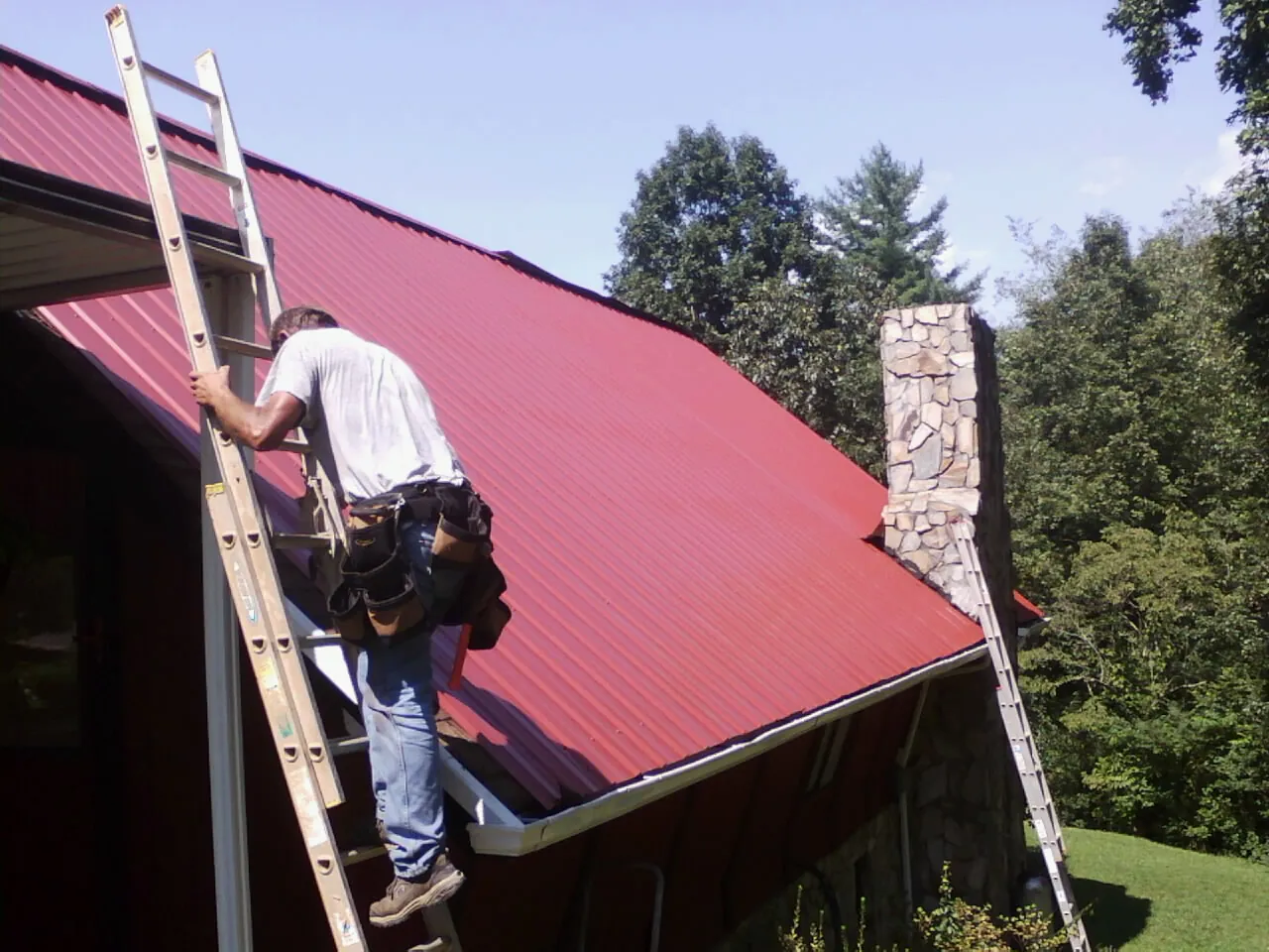 Roofer on a ladder beside a red metal roof and stone chimney — Soffit & Fascia Repair by Roofing Brothers in Alder